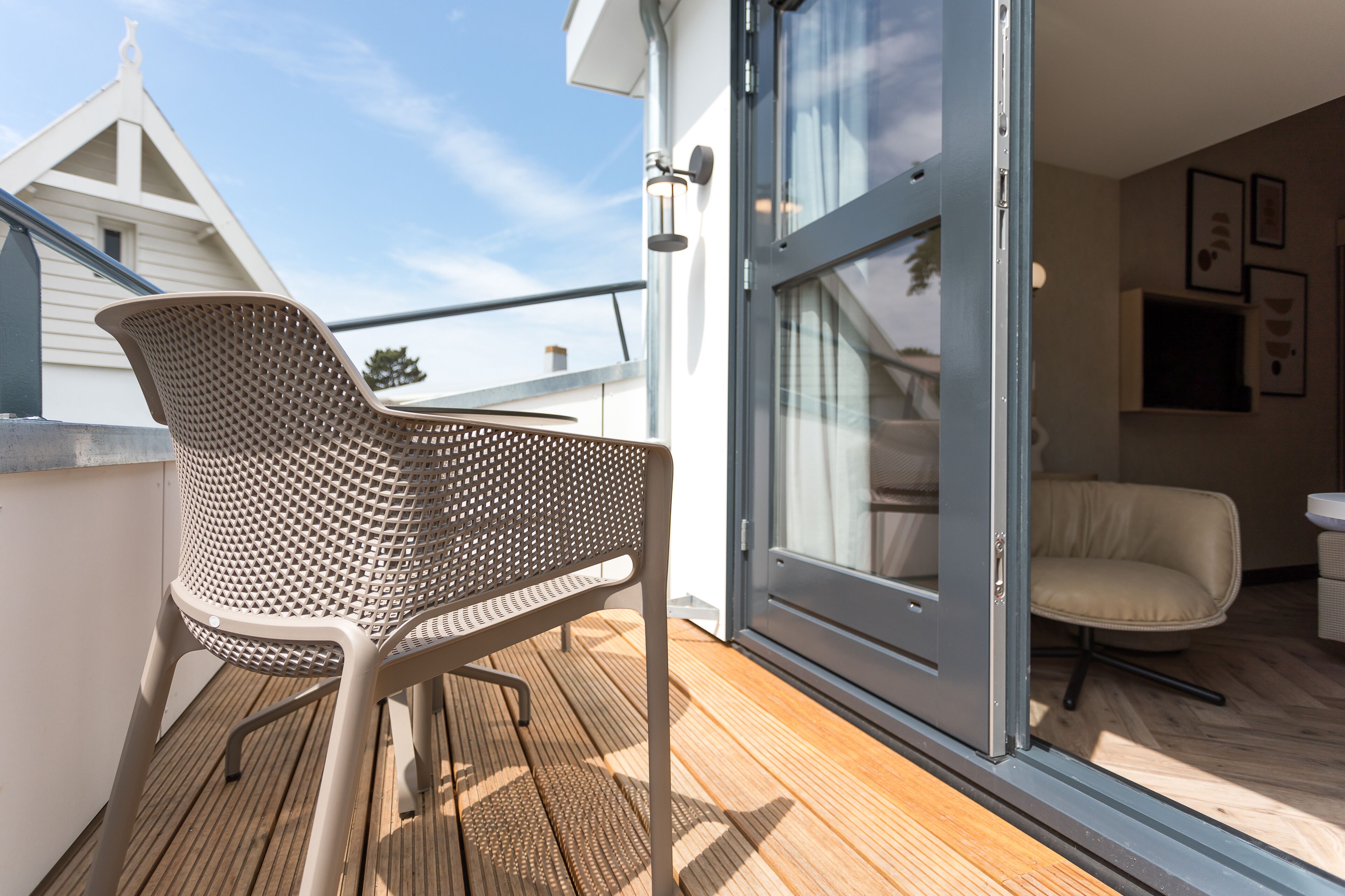 Ein Balkon mit einem Metallstuhl und einer Glastür zu einem Innenraum mit einer Couch und einem Sessel.