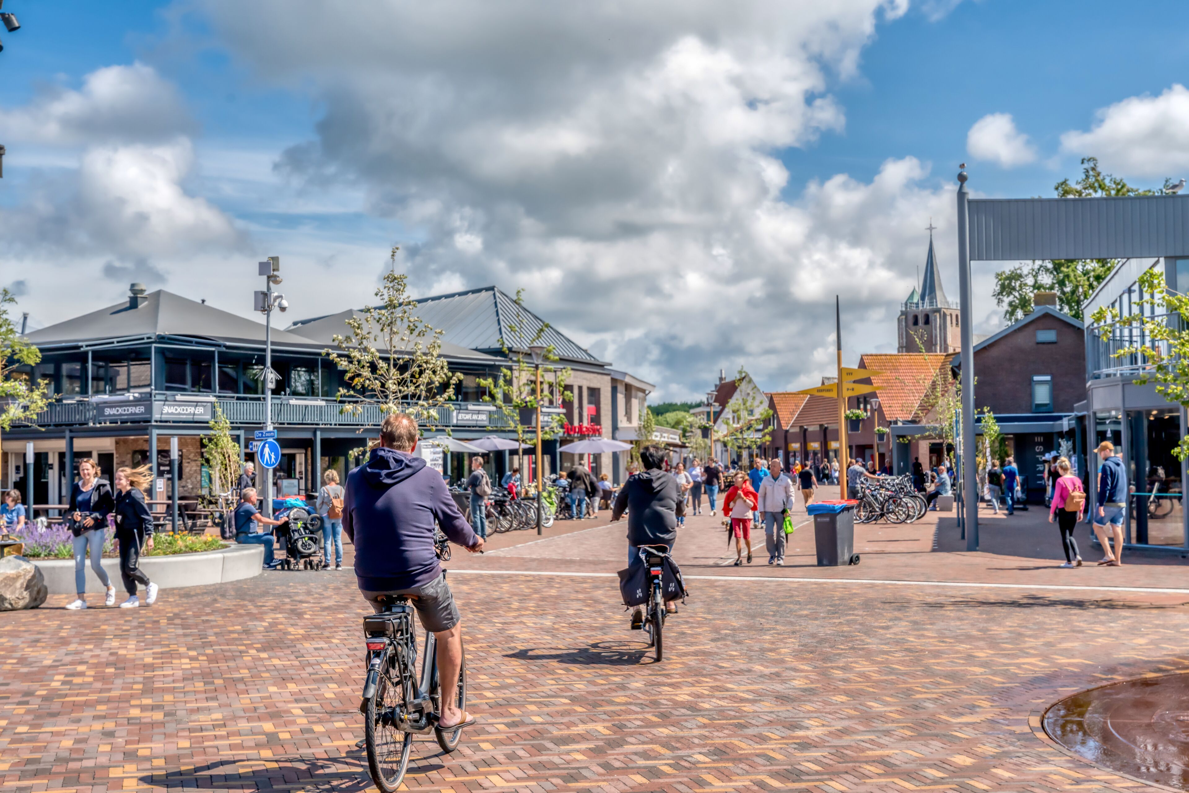 Une place publique où l’on peut voir des gens se promener et faire du vélo, entourée de bâtiments abritant divers commerces et une église.