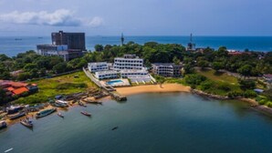 An aerial view of a coastal resort with multiple buildings, a beach, and boats docked near the shore. - The Lead Hotel (Freetown)