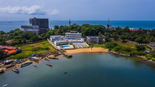 An aerial view of a coastal resort with multiple buildings, a beach, and boats docked near the shore. - The Lead Hotel (Freetown)