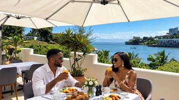 A couple dining at an outdoor table with a view of the sea and buildings in the background.