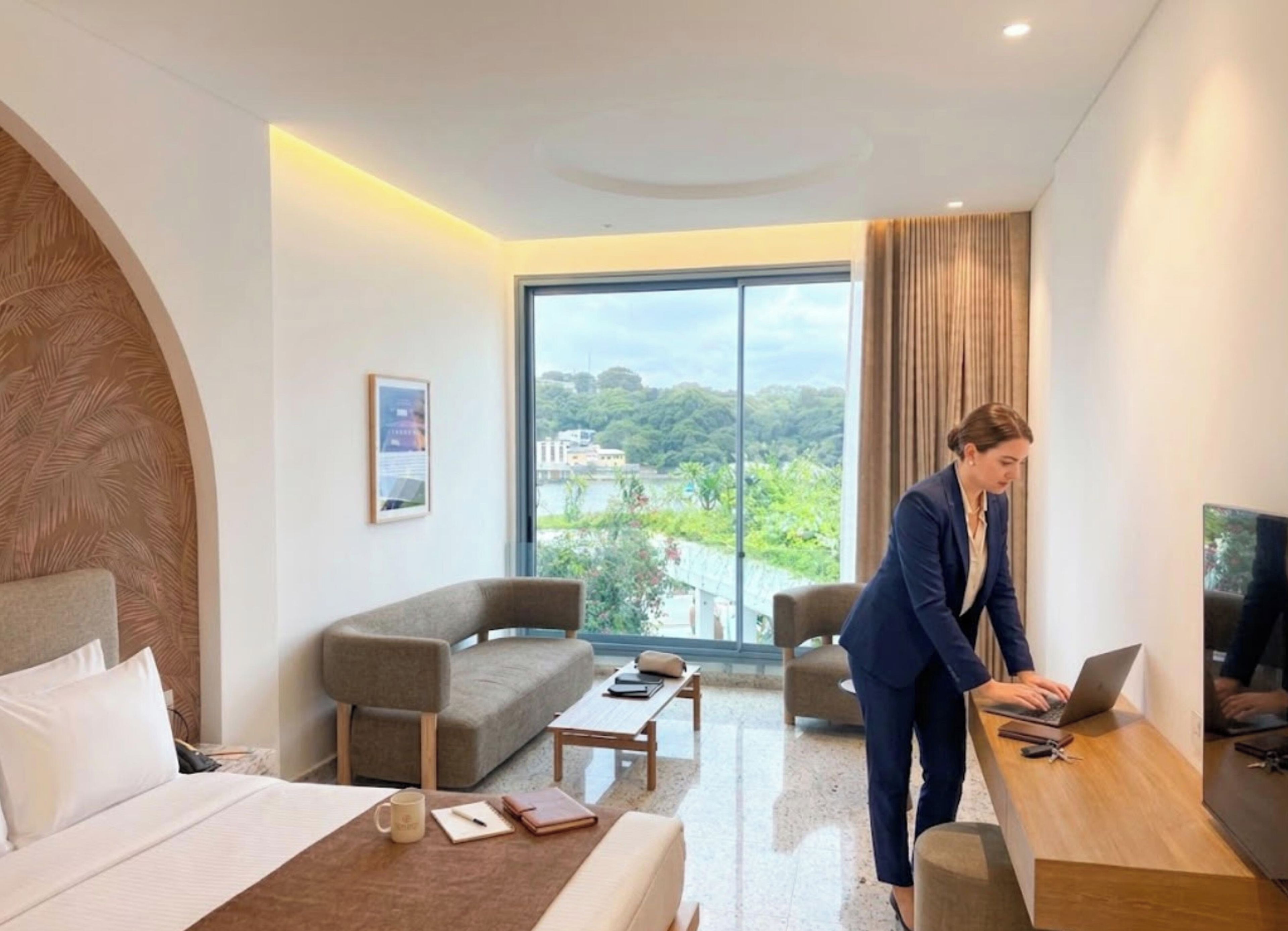 A modern hotel room with a large window, a sofa, a bed, and a desk with a laptop.