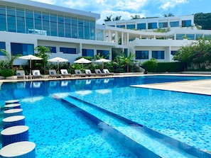 A swimming pool with lounge chairs and umbrellas, surrounded by a modern building with large windows. - The Lead Hotel (Freetown)