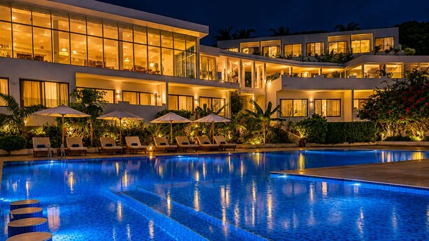 Hotel com piscina, espreguiçadeiras, guarda-sóis e fachada bem iluminada.