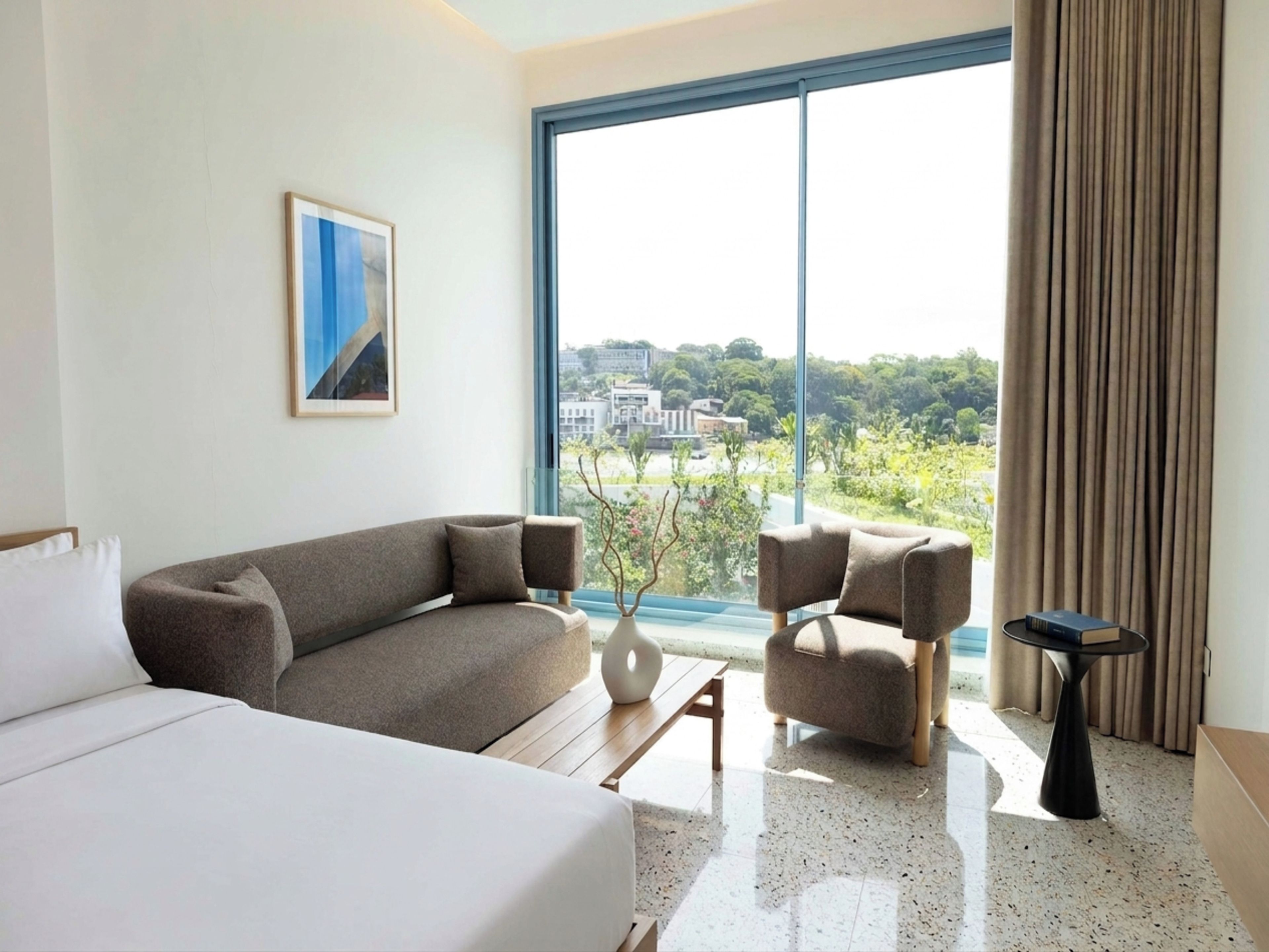 A hotel room with a bed, a sofa, a chair, a small table, and a large window with a view of buildings and greenery.