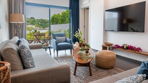 A modern living room with a sofa, armchair, and a coffee table. There is a TV mounted on the wall and a view of greenery outside. - The Lead Hotel (Freetown)