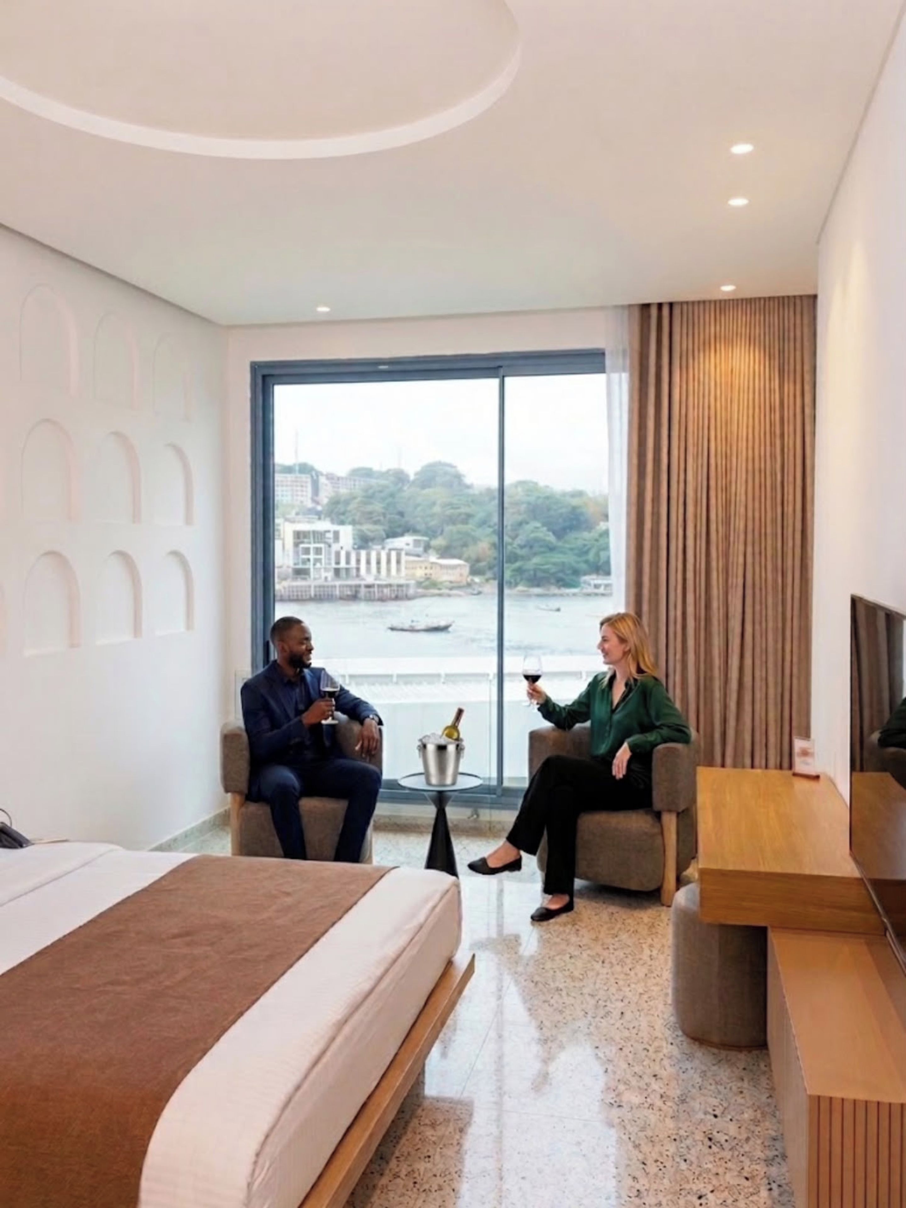 A modern hotel room with a large bed, two armchairs, a small table with a bottle, and a view of a body of water and buildings.