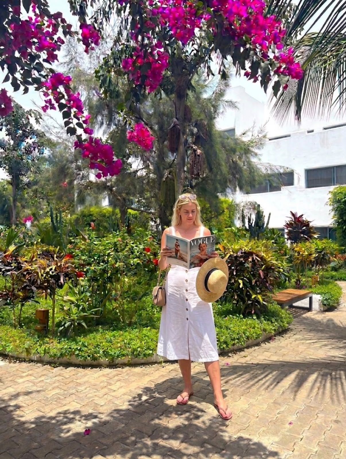 A person standing in a garden with a magazine, surrounded by lush greenery and flowering plants.