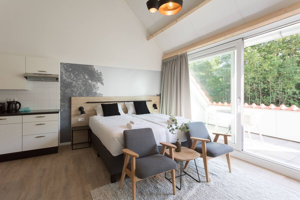 Un dormitorio moderno con una cama grande, dos mesitas de noche, una pequeña mesa con sillas, un ventanal con vistas a árboles y una zona de cocina con armarios y una cafetera.