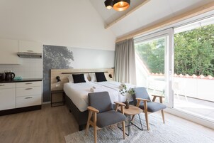 Ett modernt sovrum med en stor säng, två sängbord, ett litet bord med stolar, ett stort fönster med utsikt över träd och en köksdel med skåp och en kaffebryggare.