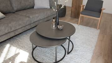 En moderne stue med en grå sofa, et rundt sofabord med to niveauer, en træstol og en vase med grene.