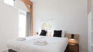 A modern bedroom with a large bed, two bedside tables, a window with curtains, and a wall-mounted light fixture. - Hofstede Moesbosch l Kloeg Collection (Koudekerke)