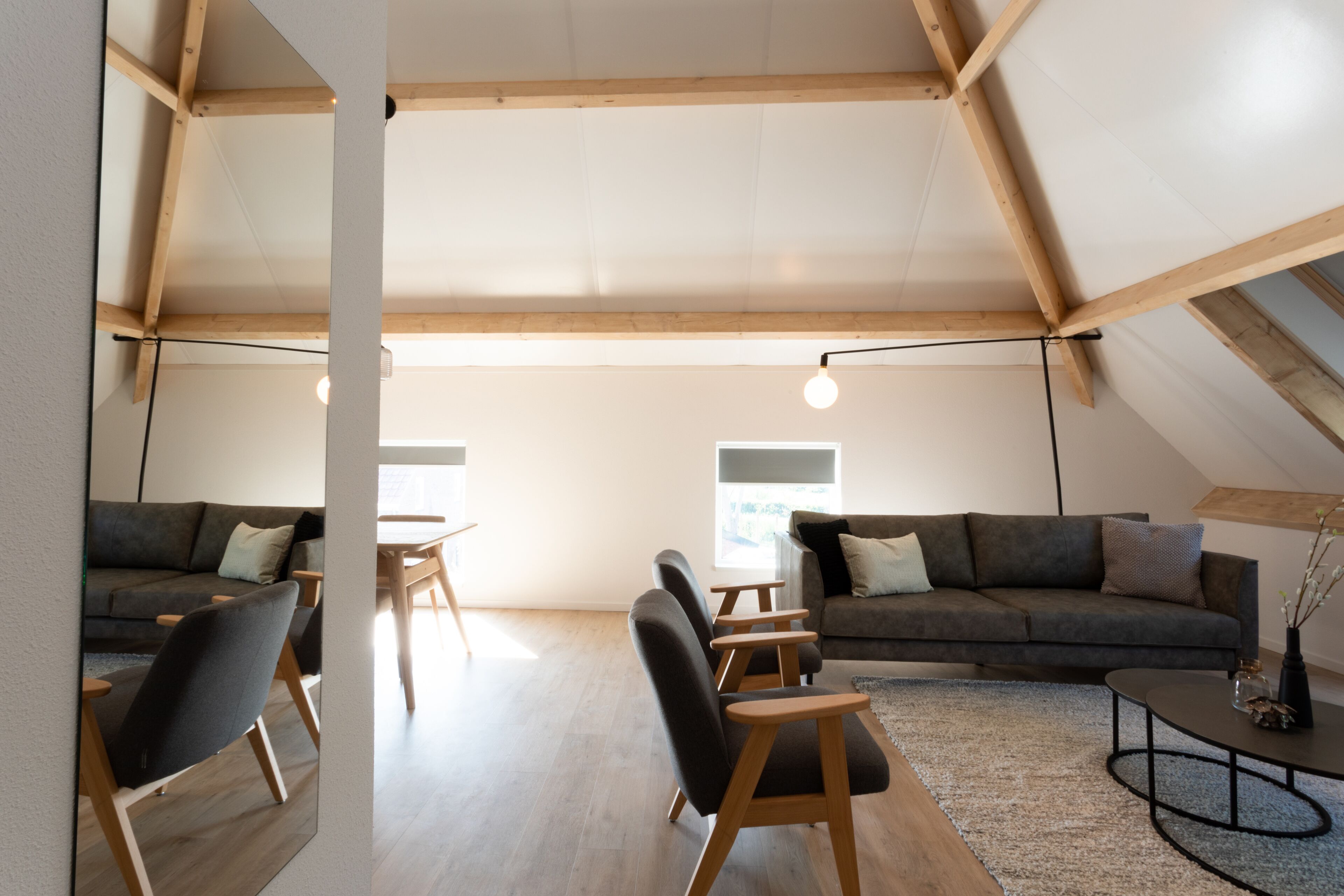 En moderne stue i loftstil med højt til loftet, synlige træbjælker, en grå modulsofa, et spisebord og et sofabord.