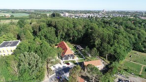 Luchtbeeld van een woonwijk met een opvallend huis met een rood dak, omgeven door weelderige begroeiing en een klein parkeerterrein.