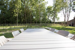 Een lange witte tafel met stoelen in een buitengebied, omringd door een hek en bomen.