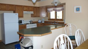 A kitchen with wooden cabinets, a refrigerator, stove, microwave, and a sink. There are two pendant lights, a window with curtains, and a trash bin. - Our Cottage Unit-Private end unit of Yellowstone Basin Inn (Gardiner)