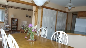 A dining area with a wooden table, white chairs, a vase with flowers, a bookshelf, and a kitchen in the background. - Our Cottage Unit-Private end unit of Yellowstone Basin Inn (Gardiner)