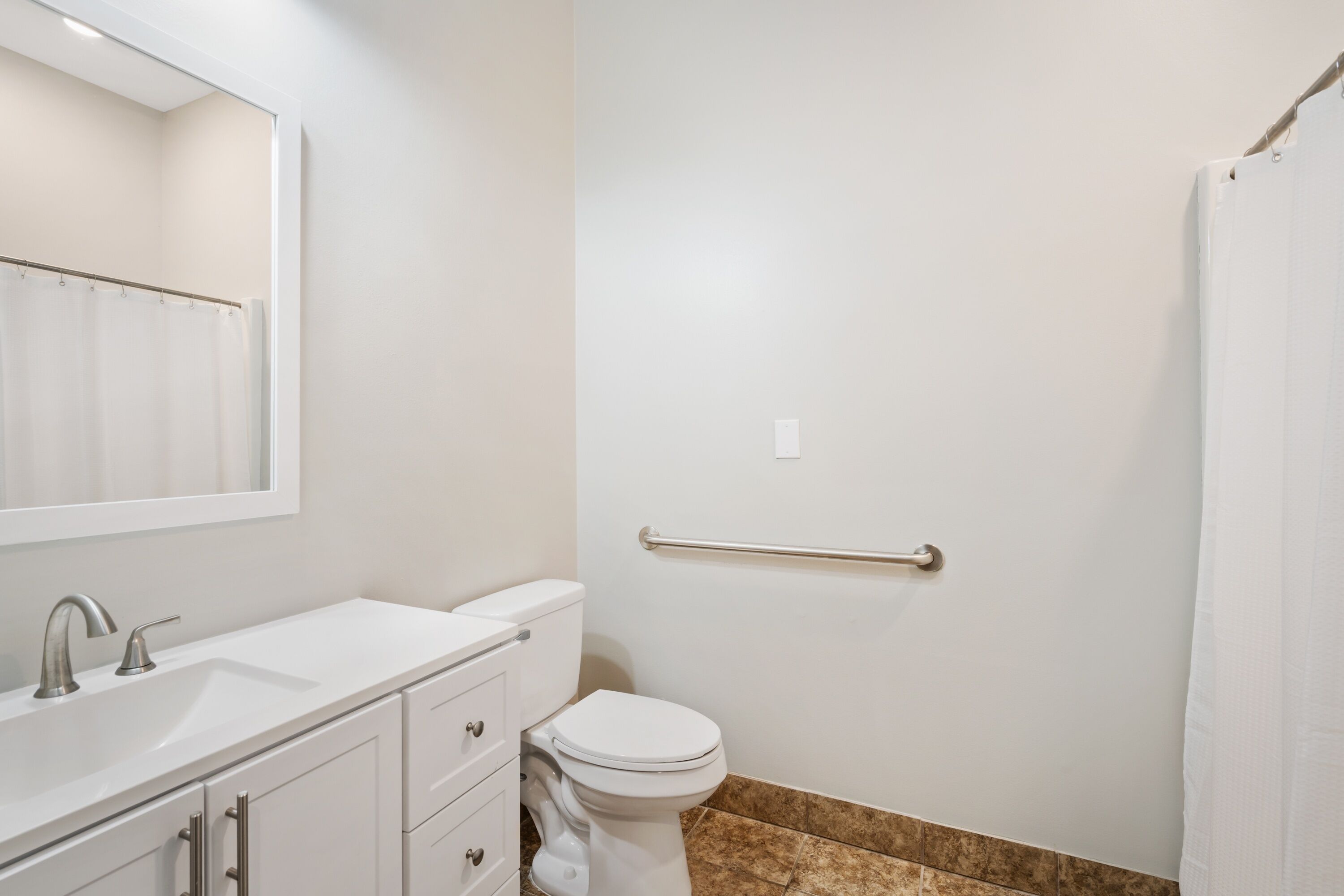 Een badkamer met een witte wasbak, spiegel en toilet. Er is een douchegordijn en een handgreep aan de muur.