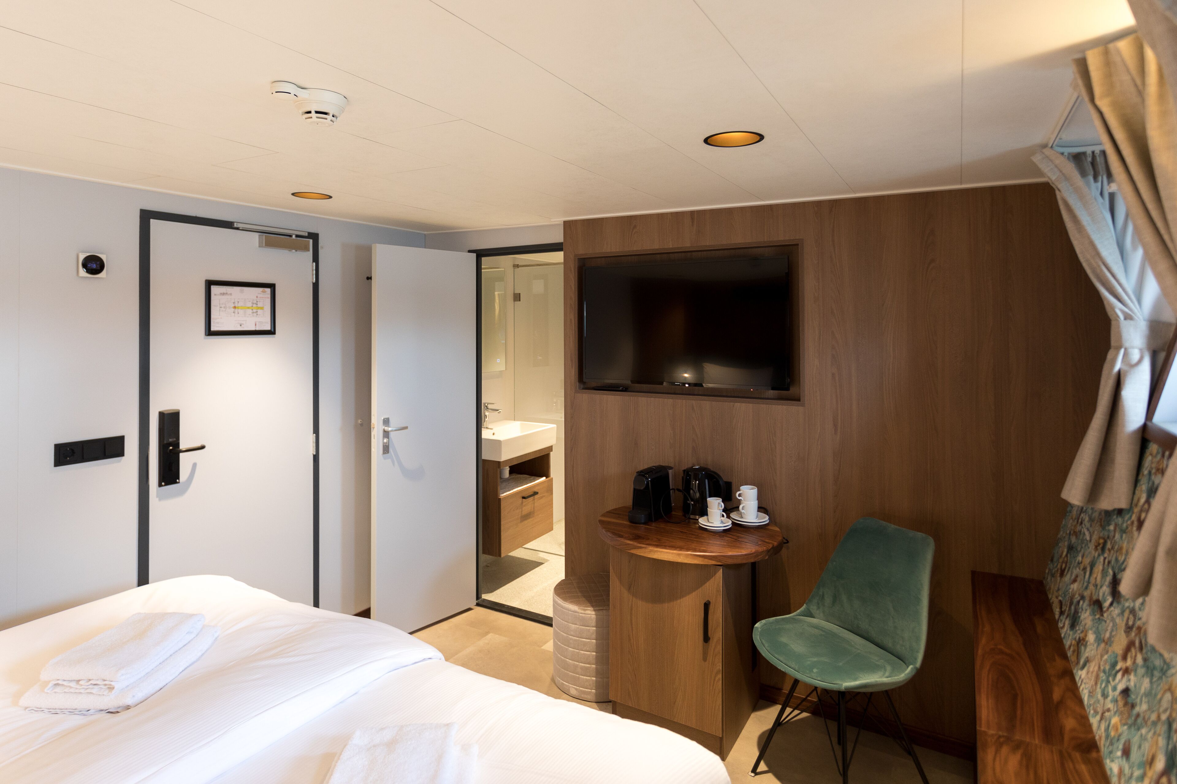 Ein Hotelzimmer mit einem Bett, einem kleinen Holztisch mit Stuhl, einem an der Wand befestigten Fernseher und einem durchgegangenen Tür erkennbaren Badezimmer.