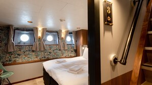 A bedroom with a bed, a chair, a window with curtains, a wall-mounted light, and a door with a number plate. - Hotelboot Koningin Emma Kloeg Collection (Vlissingen)