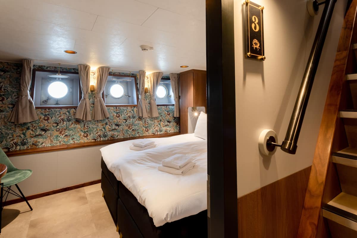 A bedroom with a bed, a chair, a window with curtains, a wall-mounted light, and a door with a number plate.