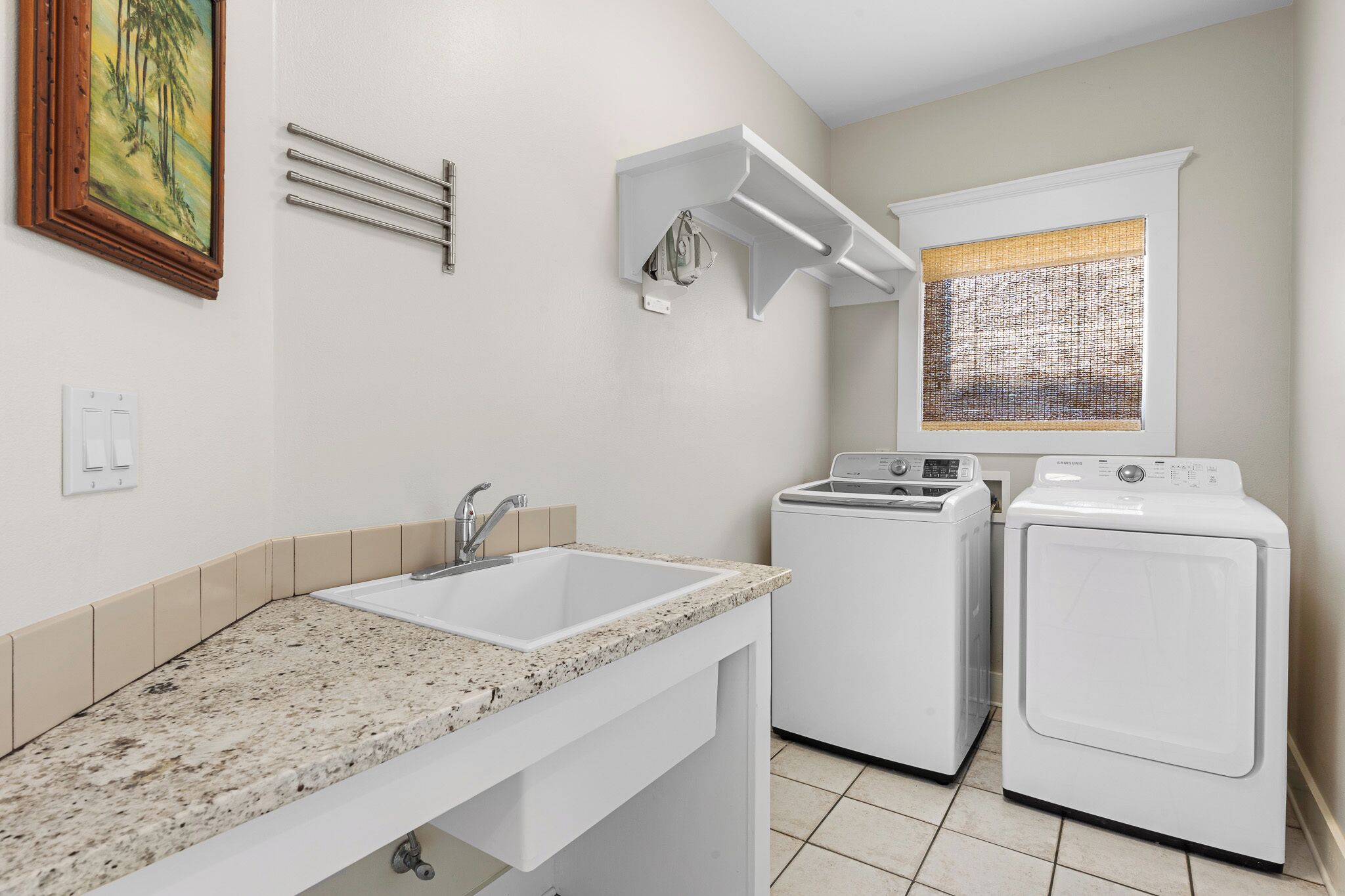 A compact laundry room with a sink, two washing machines, and a window with a textured blind.