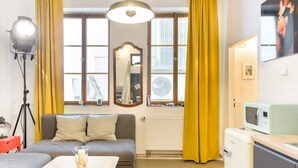 Un espacio habitable compacto con sofá, una mesita, kitchenette y un amplio ventanal con cortinas.