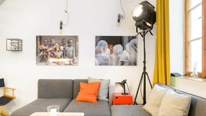 Un salón moderno con un sofá gris, cojines naranja y blanco, una mesita con un vaso y una lámpara de pie.