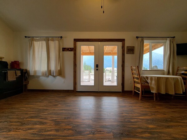 Quarto com piso de madeira, mesa com toalha, cadeira, sofá, televisão e vista para o exterior através de uma porta.