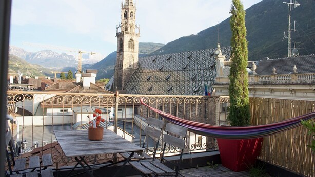 Eine Dachterrasse mit einer Hängematte, einem Tisch mit einer Topfpflanze und Blick auf eine Kirche und Berge.