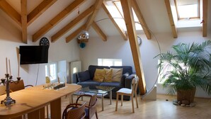 Ein gemütlicher Loft-Wohnraum mit hoher Decke, freiliegenden Holzbalken, einer bequemen Couch, einem Esstisch mit Schachspiel und einer großen Kübelpflanze.