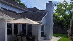 A covered patio with a table and chairs, a white umbrella, and a brick chimney. - Corporate rental, great for families in Energy Corridor. (Houston)