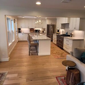 A modern kitchen with white cabinets, a central island, stainless steel appliances, and a dining area with a sofa and coffee table. - Corporate rental, great for families in Energy Corridor. (Houston)