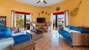 A living room with a TV, wicker furniture, and a balcony with a red railing. - Golden Zone 2/2 w Pool & Patio (5 Blocks to Beach) (Bucerías)