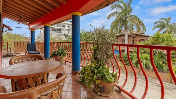 Un espace extérieur aménagé, abrité et doté de mobilier en bois, de plantes en pot, et offrant une vue sur un bâtiment et des palmiers.