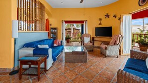 A living room with a blue sofa, wicker chairs, a TV, and a balcony with plants. - Golden Zone 2/2 w Pool & Patio (5 Blocks to Beach) (Bucerías)