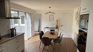 A modern kitchen with a dining area, featuring a large window, a built-in oven, and a dining table with chairs. - LE POULORIO TERRASSE - T3 RDC - 2 salles de douches  * ByLocly * (Lorient)