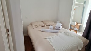 Un lit bien fait, recouvert d’une couverture blanche et agrémenté de serviettes pliées, un plancher en bois et une vue sur un balcon avec une chaise et une table.