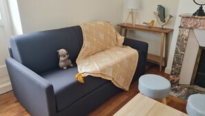 Un canapé bleu foncé, recouvert d’un plaid à motifs, avec un petit jouet en peluche et une table d’appoint en bois sur laquelle se trouvent une lampe et des oiseaux décoratifs.