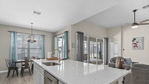 Une cuisine moderne dotée d’un grand îlot central, un coin repas avec une table et des chaises, et un salon avec un canapé et un ventilateur de plafond.