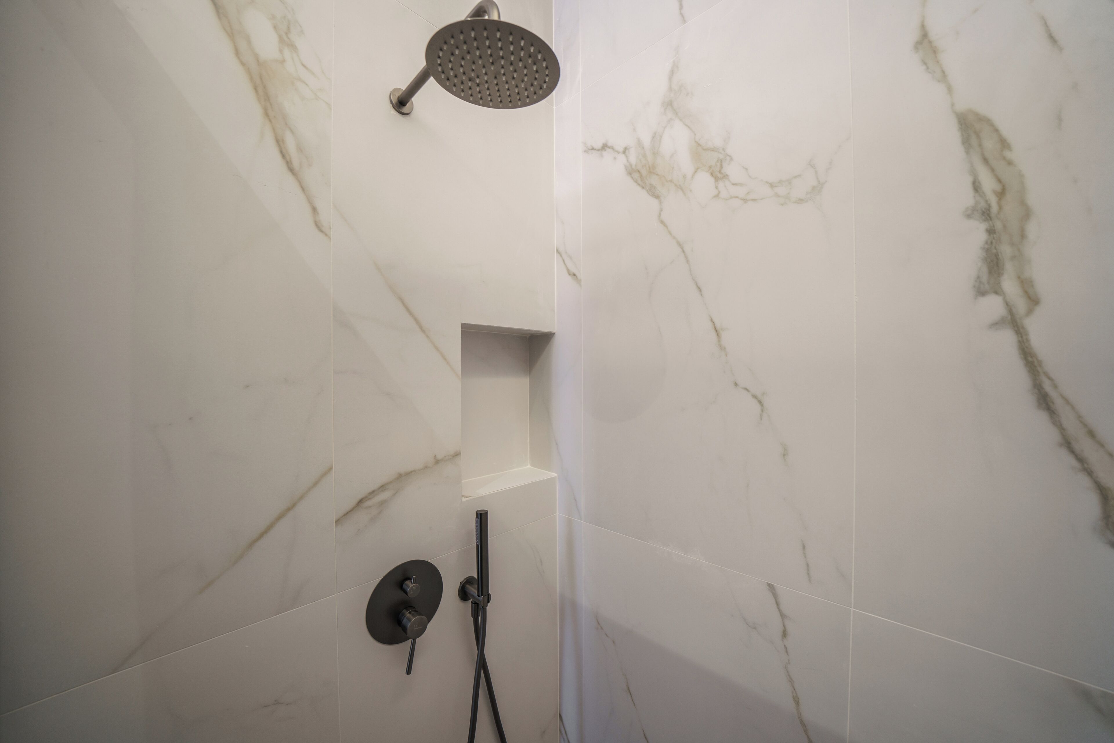A shower with a handheld showerhead, a wall-mounted control, and a built-in shelf.
