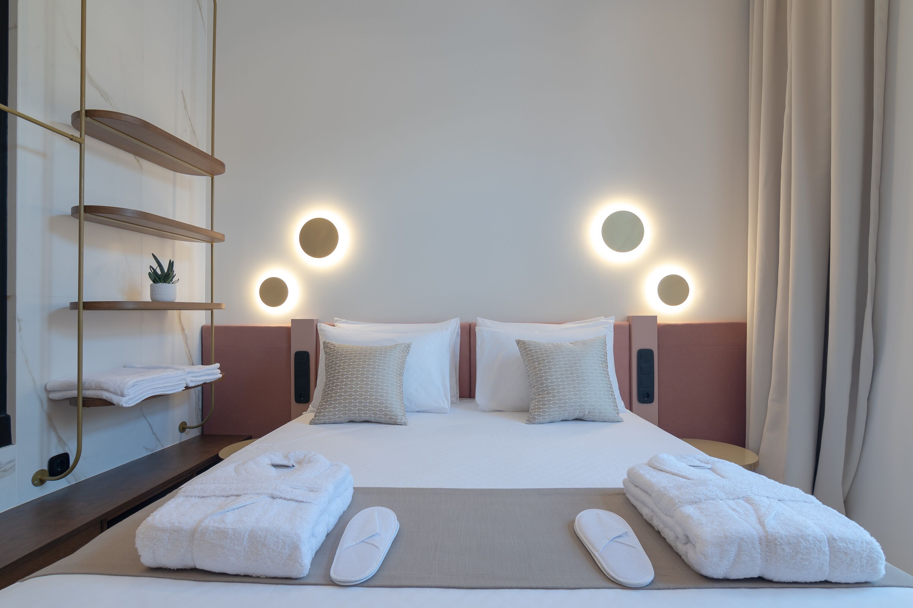 A hotel room with a bed, two bedside tables, a shelf with towels, and two wall-mounted lights.