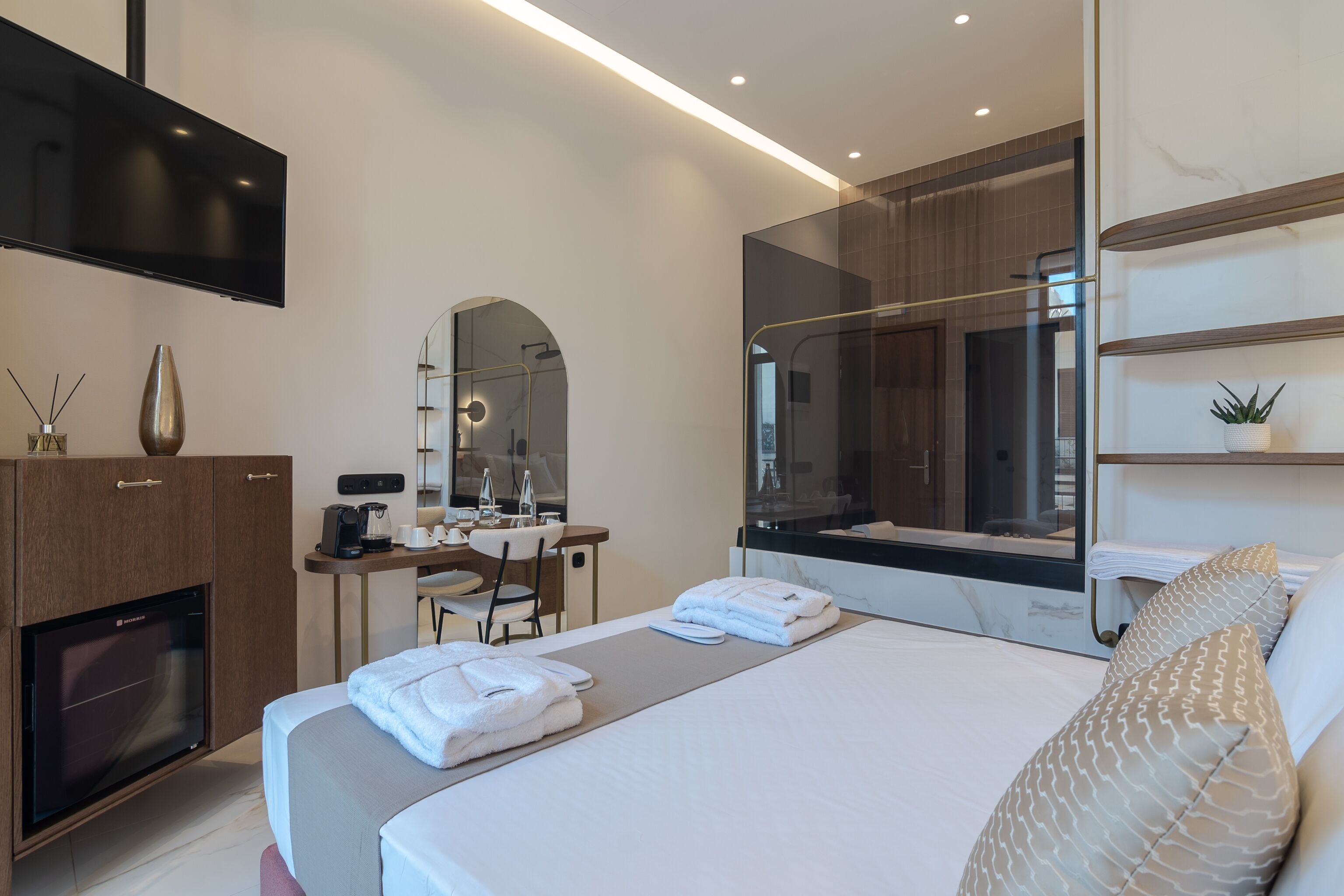 A modern hotel room with a large bed, a flat-screen TV, a desk with a chair, a mirror, and a minibar.