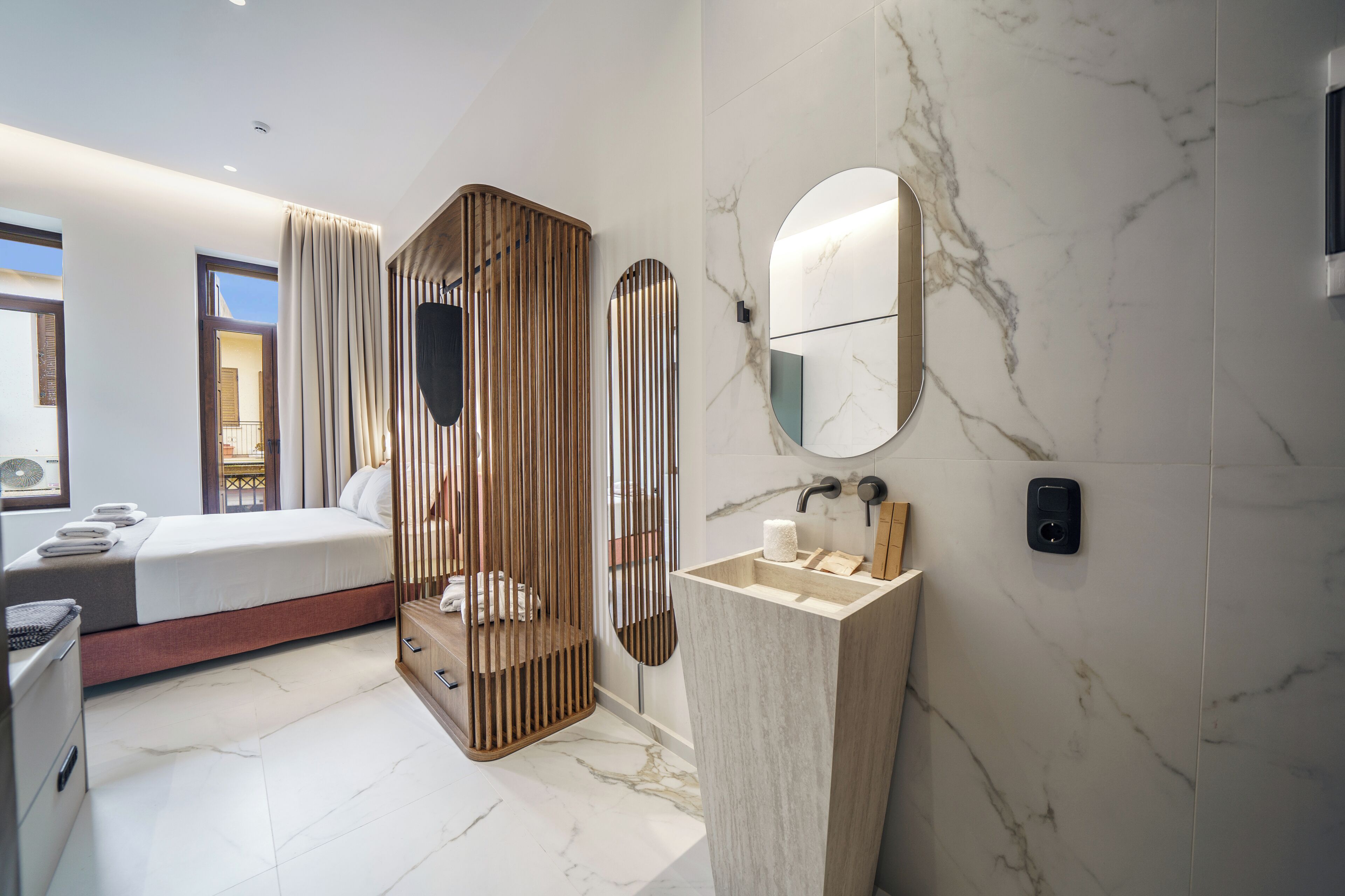 A modern hotel room with a bed, a wooden partition, a marble bathroom with a sink and mirror, and a view of the outside.