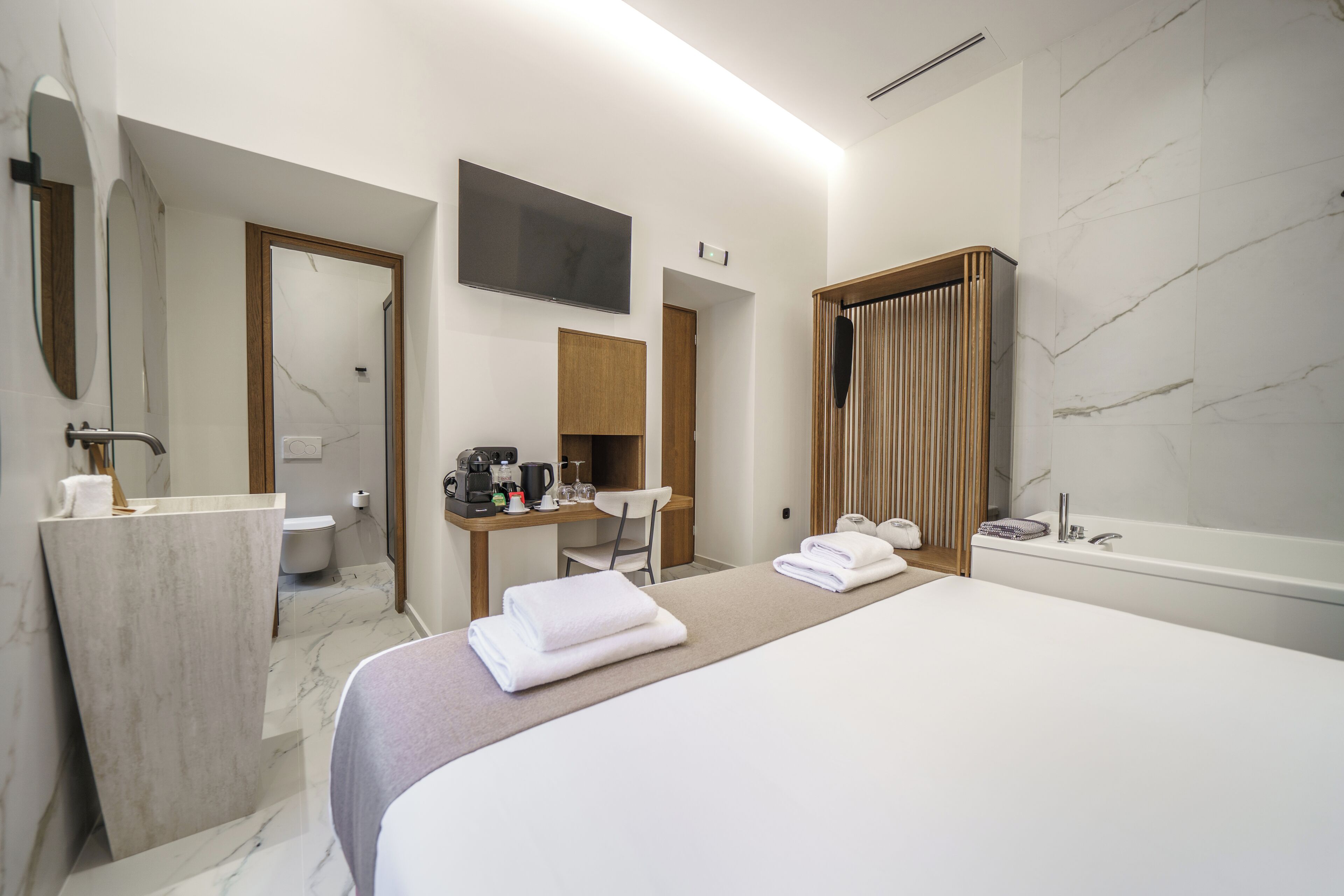 A modern hotel room with a large bed, a bathroom with a bathtub, a desk with a chair, and a flat-screen TV mounted on the wall.