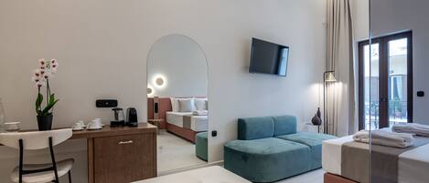 A modern hotel room with a bed, a sofa, a desk with a chair, a TV mounted on the wall, and a large mirror.