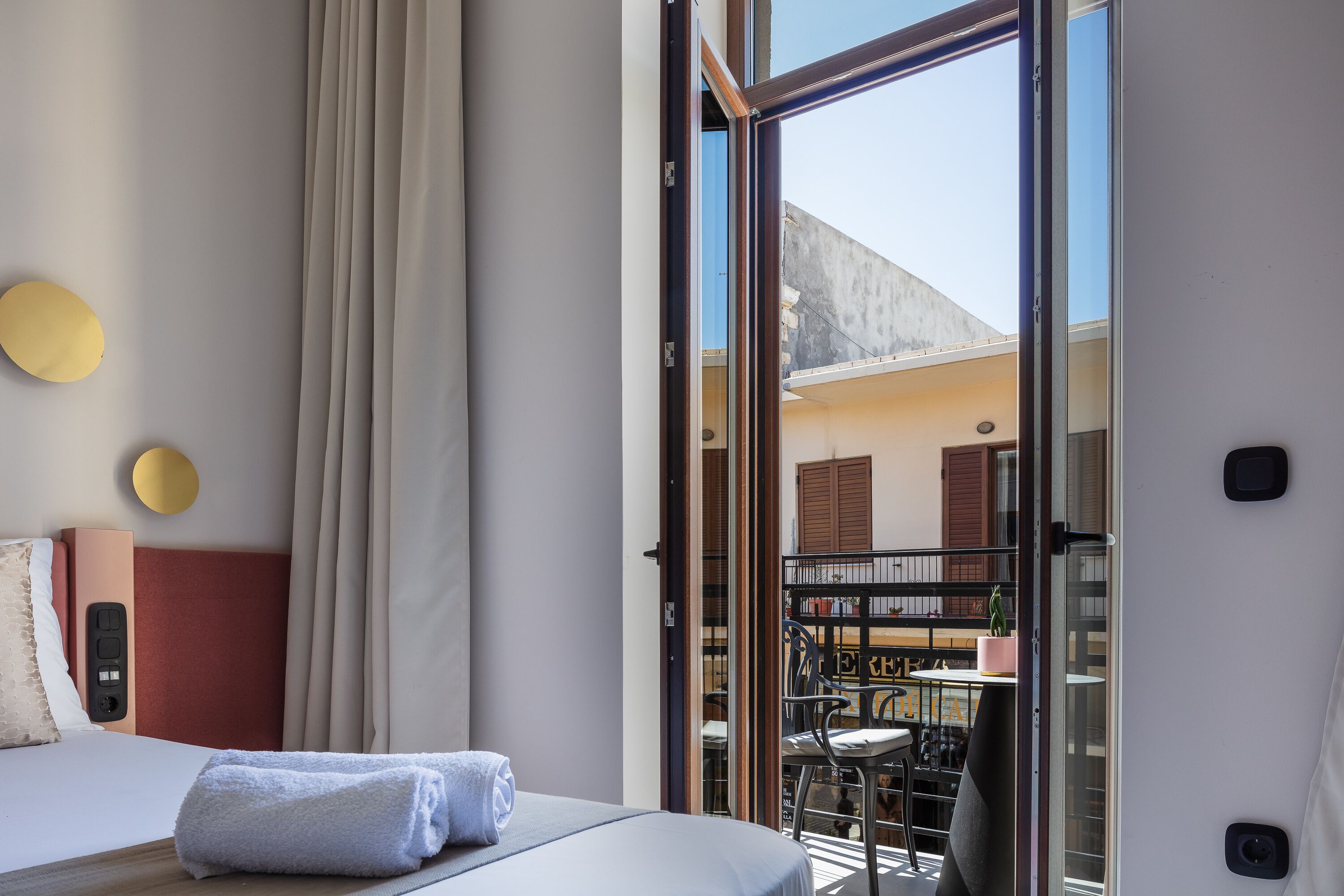 A hotel room with a bed, a bedside table, a window with curtains, and a view of the outside balcony.