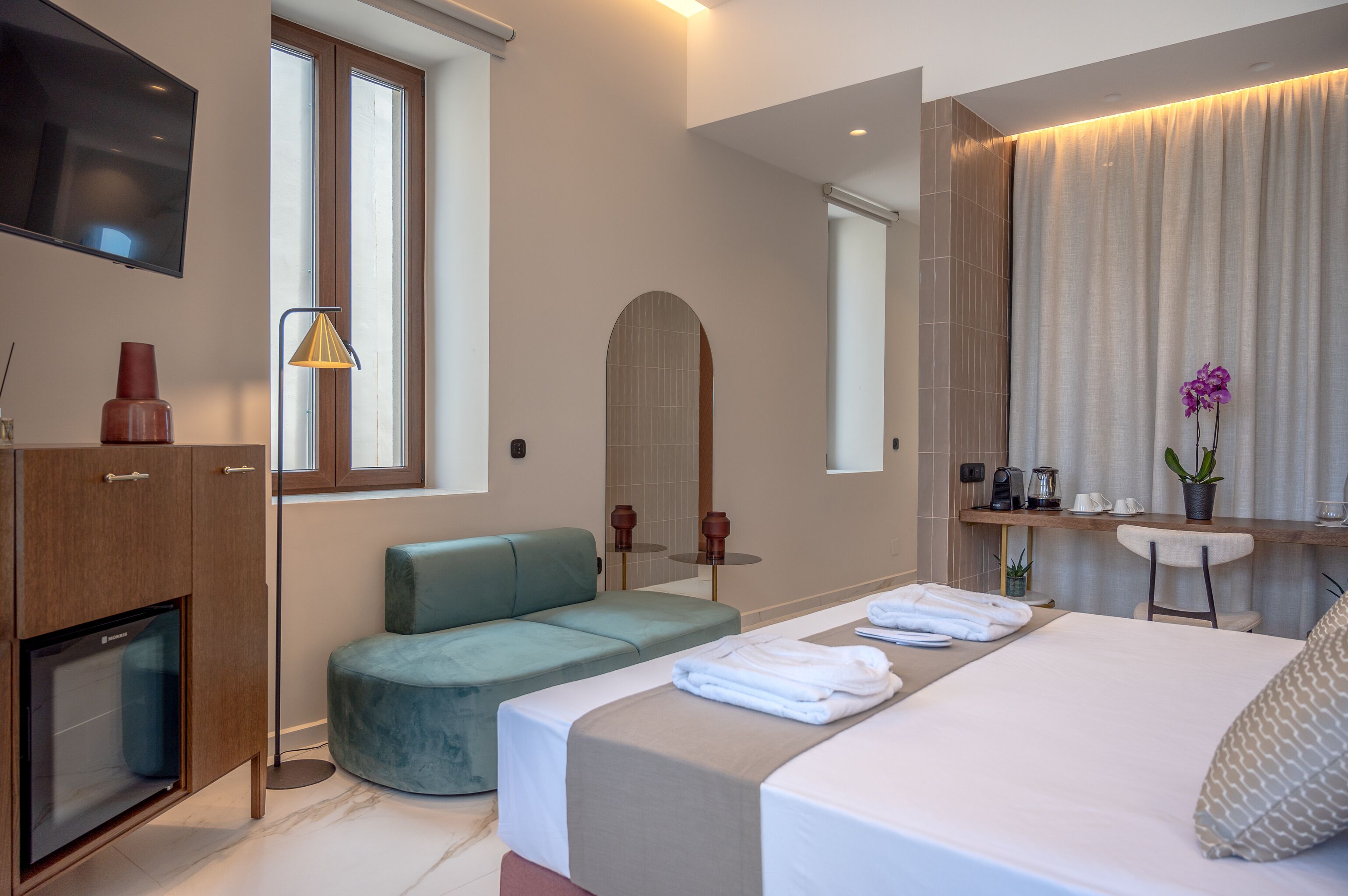 Een moderne hotelkamer met een bed, zithoek, televisie en bureau met spiegel en planten.