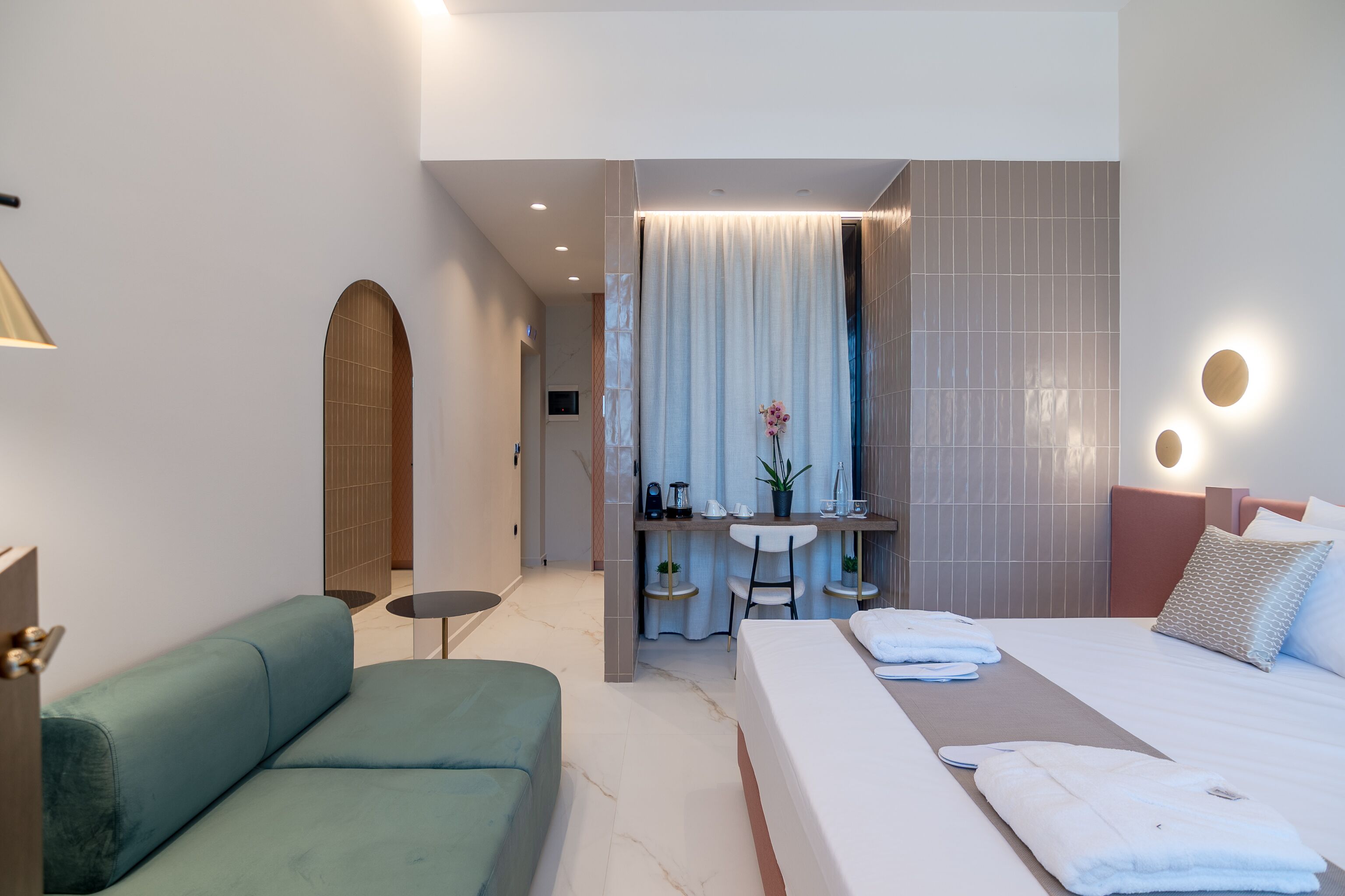 A modern hotel room with a large bed, a sofa, a desk, and a bathroom visible through an arched doorway.