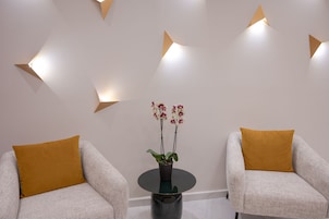 Dos sillones con cojines color mostaza, una mesita redonda con una orquídea en maceta e iluminación de pared geométrica.